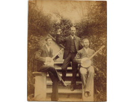 Photograph of two Banjo players seated on outdoor staircase, with another man standing playing the castanets.